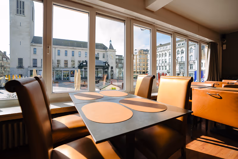 Sunlit restaurant interior with brown leather chairs and a table set with round placemats beside large windows showing historic buildings outside.