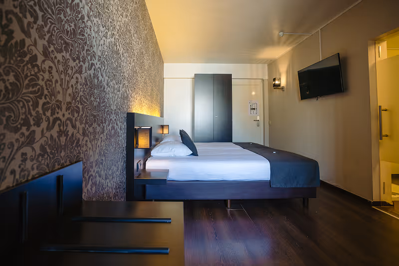 Modern hotel room with a neatly made bed, patterned wallpaper, flat-screen TV on the wall, and dim lighting.