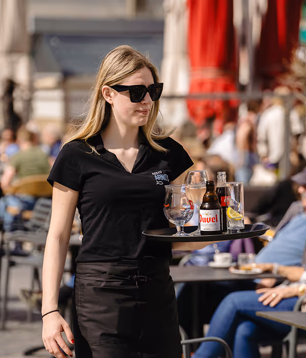 Female waitress wearing black and sunglasses carrying a tray with drinks outdoors at a busy cafe.