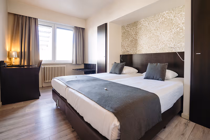 Modern hotel room with two single beds pushed together, gray bed runners, gray pillows, dark wooden headboard, patterned wallpaper behind the headboard, desk with lamp, and large window with curtains.